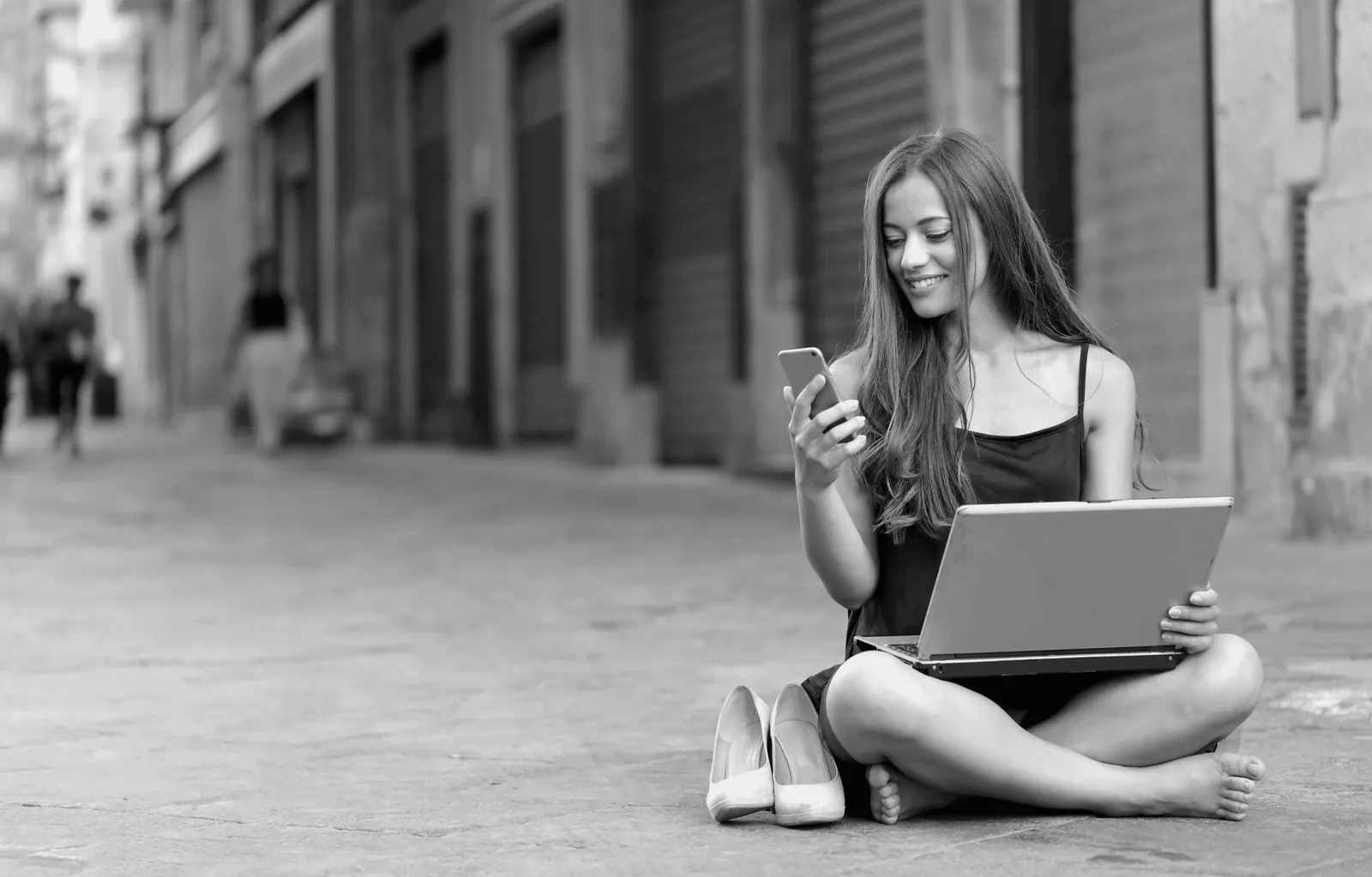 A woman sitting on a city street with a laptop, reading her phone